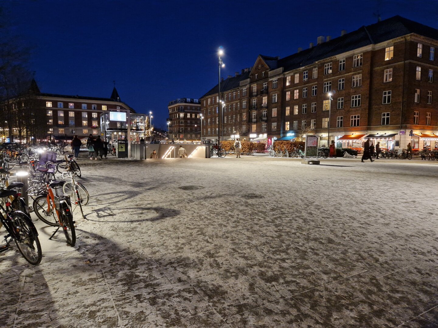 A square with a Metro exit, it is dark but with lots of street light. A thin layer of snow cover the ground.