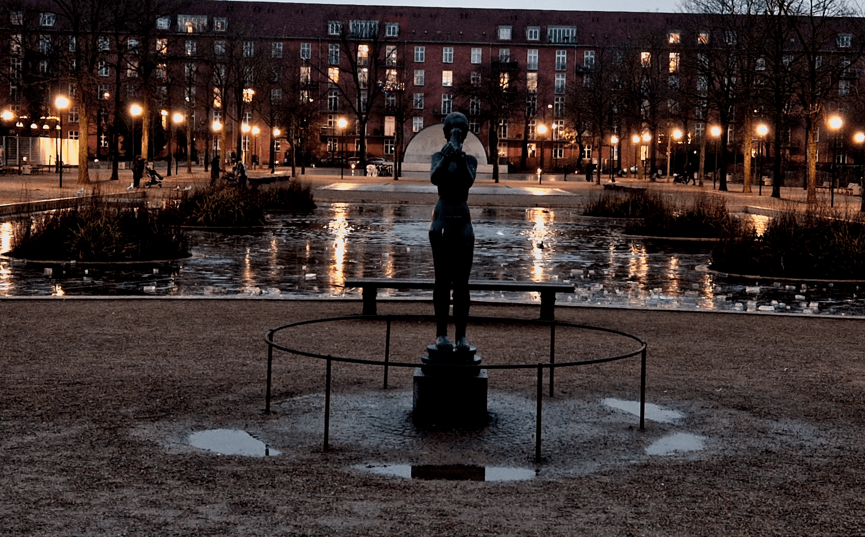 A park. A statue of a woman holding an infant in raised hands, standing in front of a pond in the background is an outdoor stage in front of a residential building.